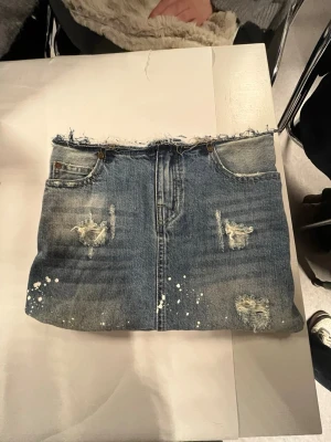 Datorfodral gjort av återvunnet jeanstyg - Säljer ett cool datorfodral med rå kant upptill, slitna detaljer och vita färgstänk. Datorfodralet har klassiska fickor fram, och insidan är fodrad med vit spets för en unik touch. Perfekt för dig som gillar att sticka ut med din stil.