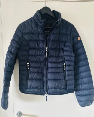 Mörkblå pufferjacka från Cedrico - Säljer en mörkblå pufferjacka från Cedrico i storlek M. Jackan har quiltad design, hög krage och två fickor med dragkedja. Stora dragkedjor framtill och snygg logga på armen. 