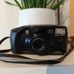 Minolta Riva Zoom 90EX - Minolta Riva Zoom 90EX är en kompakt analog kamera med autofokus och zoomobjektiv (38-90mm, f/3.5-7.7). Kameran är i gott skick med mindre ytslitage, fungerar med 35mm film och har inbyggd blixt. Perfekt för dig som vill fotografera analogt med pålitlig prestanda. Batteri behövs bytas.
