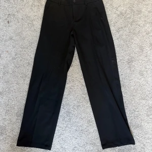 Svarta kostymbyxor Les Deux - Snygga svarta kostymbyxor från Les Deux i klassisk rak modell. Byxorna har normal passform, bälteshällor och stängs med knapp och dragkedja. Perfekta för en clean och stilren look. Materialet är mjukt och följsamt för extra komfort.