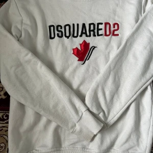 Vit Dsquared2 sweatshirt med lönnlöv - Vit sweatshirt från Dsquared2 med broderad svart och röd logga samt ett rött lönnlöv på bröstet. Tröjan har rund hals, långa ärmar och ribbade muddar. Perfekt för dig som gillar streetwear och vill sticka ut med en snygg logga.