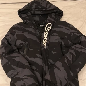 Trapstar camo pufferjacka svart/grå M - Säljer en fet pufferjacka från Trapstar i storlek M i bra skick. Jackan har svart och grått camomönster, stor huva och dragkedjor på fickorna. Trapstar-logga syns tydligt både fram och bak. Perfekt för dig som vill sticka ut i vinter. 
