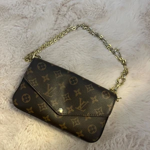  axelväska med kedja - Snygg axelväska i klassiskt monogrammönster med guldfärgad kedja. Väskan är brun och blommor, insidan är röd med plats för kort. stilren kuvertform. Inga defekt