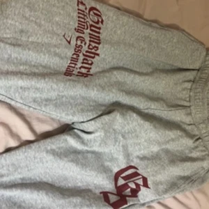 Grå mjukisbyxor från Gymshark - Grå mjukisbyxor från Gymshark med röd text 'Lifting Essentials' tryckt längs benet. Byxorna har resår i midjan och är tillverkade i mjukt bomullsmaterial. Perfekta för chill eller gymmet, med snygg logga på benet.