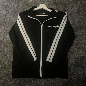 Palm Angels svart track jacket - Svart track jacket från Palm Angels med vita ränder längs ärmarna och vit dragkedja. Klassisk sportig look med logga på bröstet och ribbade muddar. Perfekt för en chill streetstyle.
