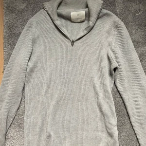Grå ribbad half zip-tröja Jack & Jones - Säljer en stilren grå ribbad tröja från Jack & Jones med half zip-dragkedja och hög krage. Tröjan är långärmad och stickad i mjukt material, perfekt för lager-på-lager under kyliga dagar.