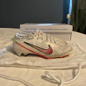 Nike Mercurial fotbollsskor Marcus Rashford - Nike Mercurial fotbollsskor i vitt med silverdetaljer och en gradient Swoosh i rött, rosa och gult. Skorna har snörning och texten 'When you believe in yourself' på sidan. Designade i samarbete med Marcus Rashford. Lätta och smidiga med platt sula och mönstrad ovandel.