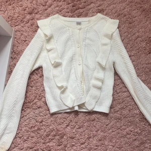 Vit kofta med volang från Lindex - Supersöt vit stickad kofta från Lindex med volangdetaljer längs axlar och fram. Koftan har rund hals, knappar framtill och ett snyggt stickat mönster. Perfekt för lager-på-lager och passar till både jeans och kjol.