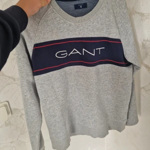 Grå sweatshirt från GANT med logga - Säljer en grå sweatshirt från GANT med marinblå panel och vit GANT-logga framtill. Tröjan har långa ärmar, rund halsringning och röda detaljer på bröstet. Tillverkad i mjuk bomullsmix, perfekt för chill dagar.