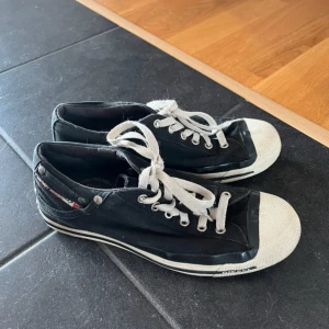 Svarta sneakers från Diesel - Snygga svarta sneakers från Diesel med vit tå och sula, klassisk låg modell med snörning och coola metalldetaljer. Skorna är i canvasmaterial och har Diesel-logga på insidan. 