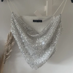 Glittrig silver topp från SHEIN - Säljer en glittrig silver topp från SHEIN i storlek S. Toppen har tunna axelband och är täckt av paljetter som ger ett riktigt snyggt partyvibe. Modellen är croppad och har en lätt draperad framsida. Perfekt för att sticka ut på nästa fest!