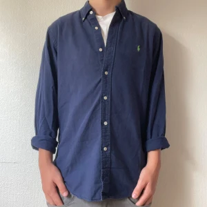 Mörkblå skjorta från Polo Ralph Lauren - Mörkblå skjorta från Polo Ralph Lauren i slim fit-modell. Skjortan har klassisk krage, knäppning framtill och en liten grön broderad logga på bröstet. Tillverkad i bomull och har långa ärmar som kan vikas upp för en mer avslappnad look. Modellen är 176cm!