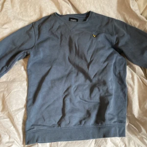 Blå sweatshirt från Lyle & Scott - Säljer en stilren blå sweatshirt från Lyle & Scott med klassisk rund halsringning och broderad gul logga på bröstet. Tröjan har långa ärmar och ribbade muddar vid ärmslut och nederkant. Perfekt för en avslappnad look.