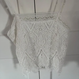 Vit spetsig linnetopp med mönster - Superfin vit linnetopp med tunna axelband och detaljerat spetsmönster. Toppen har geometriska och blommiga broderier samt en vågig kant nertill. Perfekt för dig som gillar romantisk och bohemisk stil.