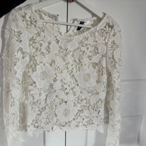 Vit spetsblus med blommor från H&M - Superfin vit blus från H&M i spets med stora blommiga broderier och pärldetaljer. Långärmad och genombruten design som ger en romantisk vibe. Perfekt för dig som vill sticka ut med en unik och detaljerad topp.