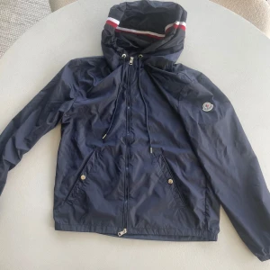 Mörkblå moncler windbreaker  - Snygg mörkblå vindjacka från Moncler och vita/röda detaljer på insidan av huvan. Klassisk Moncler-logga på ärmen och stor logotyp på insidan. Storlek 2- motsvarar ungefär M jag har S i jackor, den passar mig min lite stor. Jackan är i toppskick dessutom