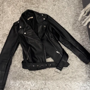 Svart skinnjacka med bälte och dragkedjor - Snygg svart skinnjacka. Jackan har silvriga detaljer, dragkedjor på fickorna och ett brett bälte i midjan. 