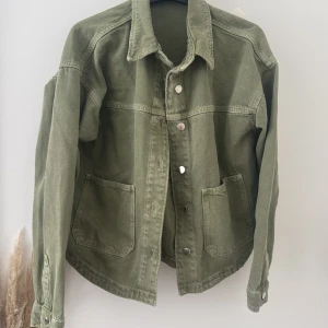 Olivgrön jeansjacka från Zara - Olivgrön jeansjacka från Zara i bomull med klassisk krage, tryckknappar framtill och två stora fickor. Jackan har raka linjer och långa ärmar med knapp vid ärmslut. 