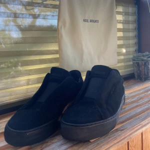 Svarta sneakers från Axel Arigato - Snygga svarta sneakers från Axel Arigato i mocka med minimalistisk design och diskret logga på sidan. Skorna har platt sula, rund tå och elastisk panel över vristen istället för klassisk snörning. Perfekta för dig som gillar stilrena och moderna skor.