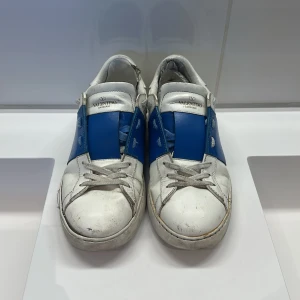 Valentino Garavani Open sneakers blå/vit - Säljer ett par välanvända Valentino Garavani Open sneakers i vitt skinn med bred blå läderdetalj över mitten. Klassisk rund tå, snörning och platt sula. Snygg och sportig design med diskreta nitar baktilll. Sulan har lossnat lite som ni ser på sista bilden, därav priset. Box och dustbag fås med!