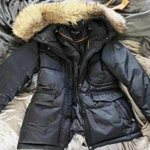 Svart Hollies dunjacka med päls - Snygg svart dunjacka från Hollies med fluffig päls på huvan. Jackan har flera fickor med lock, dragkedja och knappar framtill samt justerbar midja. Perfekt för kalla dagar med sin långa modell och varma material. Justerbar i midjan Många praktiska fickor Inga fel förutom att knapparna för pälsen är lite uttöjdas 