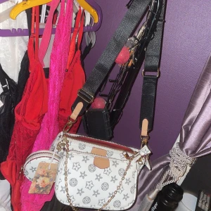 Vit axelväska med kedjedetalj - Snygg vit axelväska med svart mönster och beige detaljer. Väskan har bred svart axelrem, guldfärgad kedja och dragkedja. Tillhörande liten rund väska medföljer. Perfekt för dig som vill ha en trendig och praktisk accessoar.helt ny med pris lapp aldrig använt en 