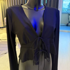 Svart croppad omlottopp med knyt - Säljer en svart croppad omlottopp med långa ärmar och djup v-ringning. Toppen har tunna ribbade ränder i tyget och knyts framtill med två smala band. Perfekt för dig som gillar trendiga och stilrena plagg.