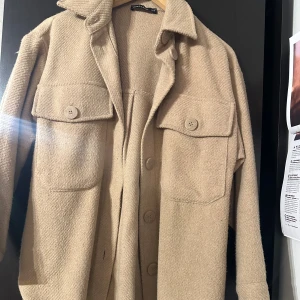 Jacka  - Stilren beige jacka i ullblandning. Jackan har stora bröstfickor med knappar, klassisk krage och knäppning framtill. Perfekt för lager-på-lager och ger en chill vibe till din outfit.