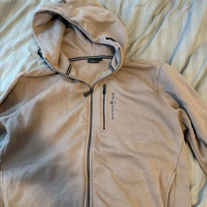 Beige zip hoddie från Sail Racing - Beige zip hoddie från Sail Racing med huva och dragsko. Jackan har hel dragkedja framtill, bröstficka med dragkedja och broderad logga. Insidan är fodrad med mjuk teddyfleece för extra värme. Perfekt för kyliga dagar.