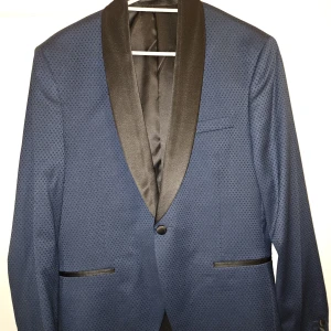 Blå kavaj med svart sjalkrage - Snygg blå kavaj med diskret prickigt mönster och svart sjalkrage i satin. Kavajen har en knapp framtill, svarta detaljer vid fickorna och en elegant, smal passform. Perfekt för dig som vill sticka ut med stilren look.