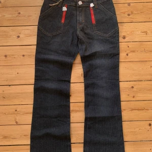 Mörkblå jeans från Ginger med röda dragkedjor W30 L30 - Unika mörkblå jeans från Ginger med raka ben och snygga kontrastsömmar. Framtill finns två röda dragkedjor som ger en cool detalj. Klassisk femficksmodell och normal passform. Perfekta för dig som vill sticka ut lite extra. Storlek W30 L30