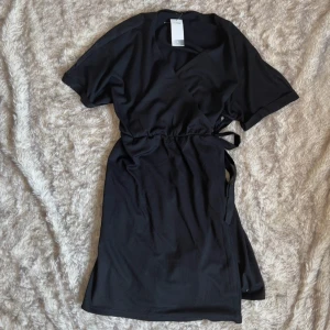 Svart omlottklänning med knytband - Svart omlottklänning  i storlek L. Klänningen har korta ärmar, justerbara knytband i midjan och en v-ringad omlottfront. Ny med prislapp