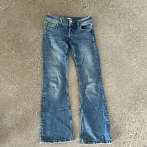 Blå bootcut jeans med dubbla knappar - Snygga blå jeans med bootcut passform och dubbla silverfärgade knappar i midjan. Jeansen har klassiska fickor fram och bak, där bakfickorna har lock och knappdetalj. Materialet är jeans i bomull och de har en lätt tvättad look.