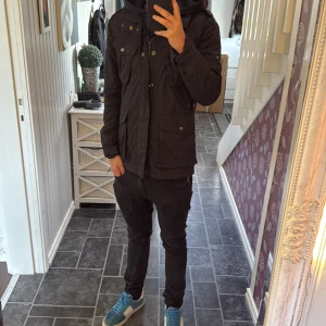 Woolrich fieldjacka  - Svart parkajacka från Woolrich med flera fickor framtill, huva och snygga detaljer i metall. Jackan har dragkedja och knappar, samt justerbar midja. Perfekt för dig som vill ha en stilren och funktionell jacka med streetkänsla. Sitter bra på mig som är 170