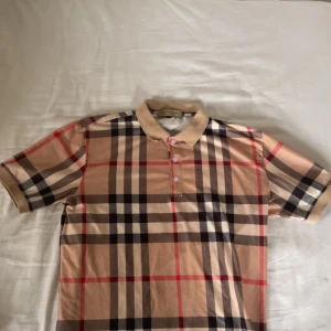 Rutig Burberry t-shirt med krage - Snygg t-shirt från Burberry med klassiskt rutig mönster i beige, svart, röd och vit. T-shirten har korta ärmar, krage och tre knappar framtill. Perfekt för dig som gillar ikoniska designer och vill sticka ut med stil.