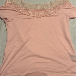 Ljusrosa topp med spetsdetalj - Supersöt ljusrosa topp med korta ärmar och bred v-ringning dekorerad med romantisk spets i matchande ton. Perfekt för dig som gillar en feminin och mjuk stil. Tillverkad i mjukt material som känns skönt mot huden.