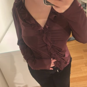 Vinröd omlottopp med volang - Säljer en vinröd omlottopp med långa ärmar och snygga volangdetaljer längs v-ringningen och framtill. Toppens material är mjukt och stretchigt, vilket gör den bekväm och smickrande. Perfekt för dig som gillar feminina detaljer och en trendig look.