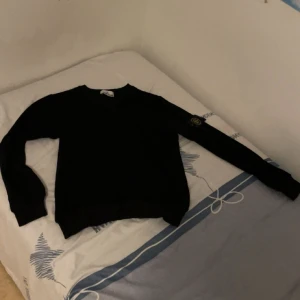 Svart Stone Island tröja i bomull - Svart långärmad tröja från Stone Island med klassisk patch på vänster ärm. Tröjan är i mjuk bomull och har en enkel, clean design med rund hals. Perfekt för dig som gillar streetwear och vill ha något stilrent med ikonisk logga. Skick 10/10 och pris går att diskutera, Om ni har frågor är det bara fråga. 