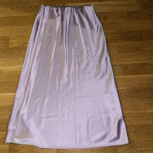 Ljuslila satinkjol från Gina Tricot - Säljer en lång, glansig ljuslila kjol från Gina Tricot i satinliknande material. Kjolen har resår i midjan och en rak, flowy passform som ger en snygg siluett. Perfekt för dig som vill ha en stilren och trendig look.