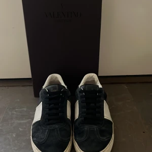 Valentino Garavani sneakers marinblå/vit - Snygga Valentino Garavani sneakers i marinblå mocka och vitt skinn med blåa snören och rund tå. Yttersulan har ett coolt camouflage-mönster och platt sula. Märkeslogga på plösen och insidan. Perfekt för dig som vill ha en lyxig look. Storlek 39 men sitter som 40-41.