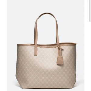 Beige väska - Stilren shoppingväska från Glitter i beige med diskret mönster av logotyper över hela väskan. Väskan har två långa handtag och detaljer i matchande beige. Perfekt för dig som vill ha en rymlig och snygg väska till vardagen.