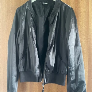 Svart bomberjacka med glansig finish - Svart bomberjacka från H&M med glansig yta och ribbade muddar. Jackan har dragkedja framtill och snörning i ryggen för en snygg detalj. Långärmad och kortare modell som ger en cool och modern look.