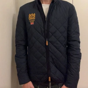 Mörkblå quiltad jacka från Morris - Snygg mörkblå quiltad jacka från Morris med guldbroderad logga och brittisk flagga på bröstet. Köpt helt ny för 2000 kr. Jackan har dragkedja, ståkrage i manchester och klassiskt rutmönster. Perfekt för dig som vill ha en stilren och tidlös look. Skriv vid minsta fundering, pris kan diskuteras!