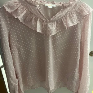 Ljusrosa blus med volang från Gina Tricot - Supersöt ljusrosa blus från Gina Tricot med prickigt mönster och volangdetalj längs ringningen. Blusen är tunn och luftig med långa ärmar.Perfekt för dig som gillar en feminin och trendig stil.