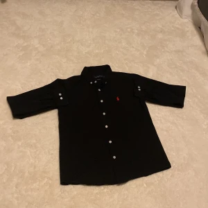 Svart skjorta från Polo Ralph Lauren - Svart skjorta från Polo Ralph Lauren med klassisk krage, vita knappar och röd broderad logga på bröstet. Skjortan har lång ärm med knappdetaljer och är tillverkad i bomull för en skön känsla. Perfekt för dig som gillar stilrena och tidlösa plagg.