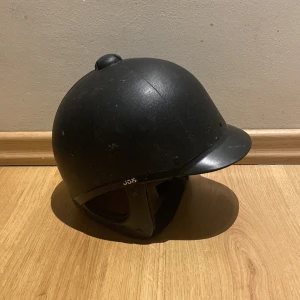 Svart ridhjälm med hakrem - Säljer en klassisk svart ridhjälm med rundad form och kort skärm framtill. Hjälmen har justerbar hakrem och ventilerad, ljusgrå insida för extra komfort. Perfekt för dig som vill ha en stilren och säker hjälm till ridningen. Vet inte om storlek på den men ungefär för en 8-11 åring. 