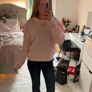 Ljusrosa stickad tröja med volang - Superfin ljusrosa stickad tröja med volangdetaljer på axlarna. Tröjan har rund halsringning och långa ärmar, perfekt för att ge en söt och trendig look. Mjuk och skön att ha på sig, passar perfekt till jeans eller kjol.