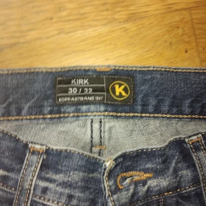 Blå jeans från Koppartrans Kirk 30/32 - Säljer ett par klassiska blå jeans från Koppartrans, modell Kirk. Jeansen har snygga slitningar, fem fickor och kontrastsömmar. Märkespatch i läder baktill och coola detaljer vid fickorna. Perfekt för dig som gillar en tidlös denimlook.