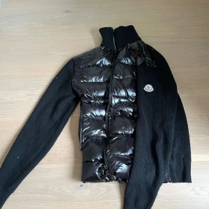 Svart puffer cardigan jacka från Moncler - Svart cardigan jacka från Moncler med glansig pufferfront och stickade ärmar. Jackan har hög krage, dragkedja och Moncler-logga på ärmen. Materialmix av dunfylld polyester och stickad ull ger både värme och stil. Perfekt för kyliga dagar.