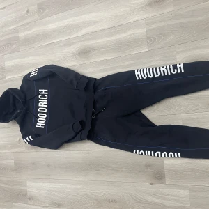 Svart Hoodrich dress - Svart Hoodrich dress med vit logga längs benet och blå piping längs sidorna. Byxorna har dragsko i midjan och är tillverkade i mjukt material som passar perfekt för chill eller streetwear. Snygg och avslappnad passform.